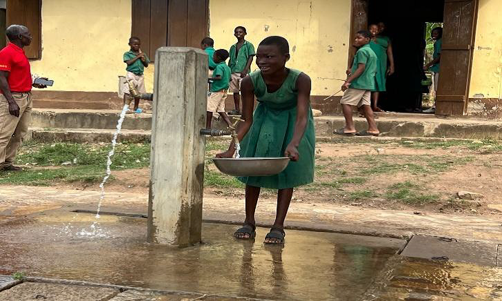 A smiling young schoolgirl in a green uniform fills a metal bowl with clean water from a concrete standpipe in a schoolyard, with other students in the background.