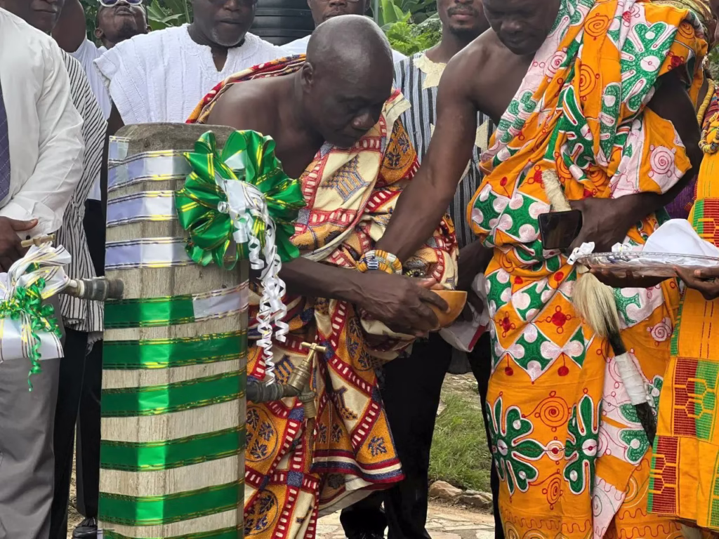 A Ghanaian traditional leader in vibrant yellow and red Kente cloth bends over to bless a new community water tap decorated with a green and white ribbon.